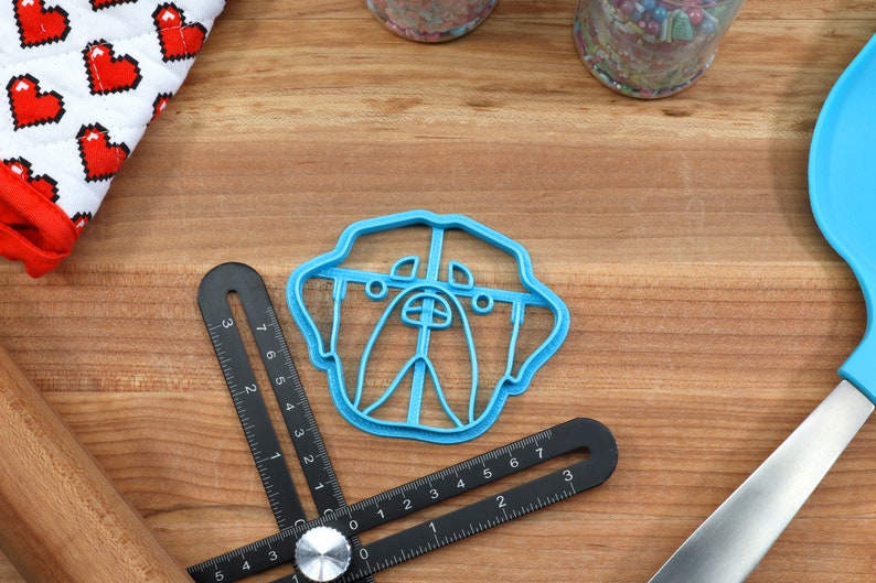May include: A blue plastic cookie cutter shaped like a dog's face. The cutter is on a wooden surface with a ruler and a rolling pin.
