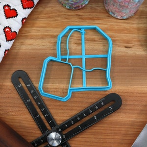 May include: A blue plastic tea bag cookie cutter on a wooden surface. The cutter is shaped like a tea bag with a string and tag.