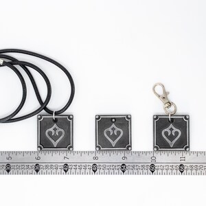 May include: Three silver-colored metal pendants with a black and white design. The pendants are square-shaped and feature a stylized heart design. The pendants are shown on a ruler with the largest pendant measuring 1.5 inches wide. The pendants are attached to a black cord and a silver key chain.