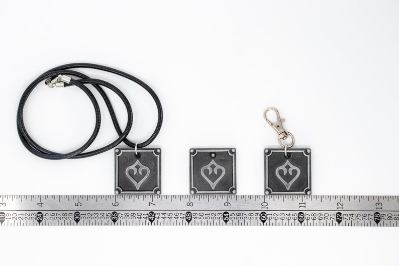 May include: Three silver-colored metal pendants with a black and white design. The pendants are square-shaped and feature a stylized heart design. The pendants are shown on a ruler with the largest pendant measuring 1.5 inches wide. The pendants are attached to a black cord and a silver key chain.