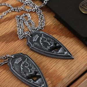 May include: Two silver necklaces with a shield-shaped pendant featuring a unicorn design. The pendants are on a wooden surface with a black leather box in the background.