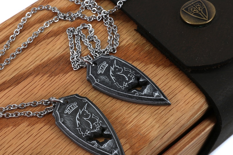 May include: Two silver necklaces with a shield-shaped pendant featuring a unicorn design. The pendants are on a wooden surface with a black leather box in the background.