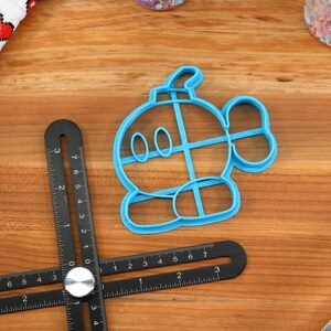 May include: A blue plastic cookie cutter shaped like a cartoon character with a round body, two eyes, and a small tail. The cookie cutter is on a wooden surface.