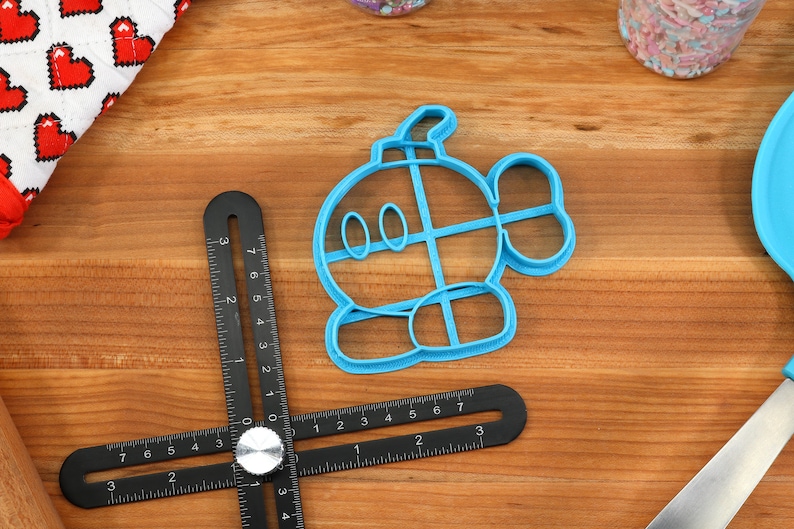 May include: A blue plastic cookie cutter shaped like a cartoon character with a round body, two eyes, and a small tail. The cookie cutter is on a wooden surface.