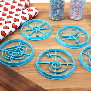 May include: Five blue plastic cookie cutters with different designs on a wooden surface. The designs include a spiral, a circle with a cross, a circle with a star, a circle with a swirl, and a circle with a flower.