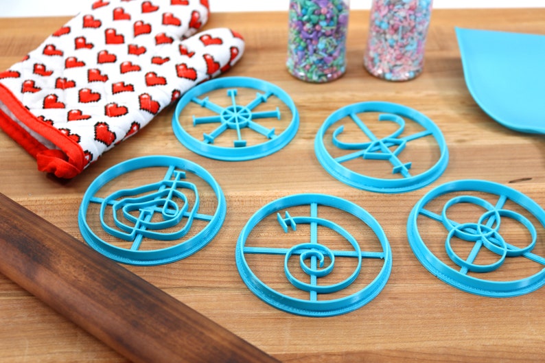 May include: Five blue plastic cookie cutters with different designs on a wooden surface. The designs include a spiral, a circle with a cross, a circle with a star, a circle with a swirl, and a circle with a flower.