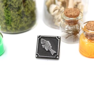 May include: A silver metal pin with a black background and a silver fish design. The pin is surrounded by a silver frame with four decorative studs.
