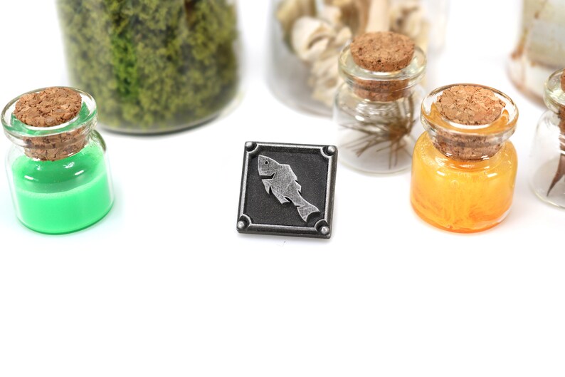 May include: A silver metal pin with a black background and a silver fish design. The pin is surrounded by a silver frame with four decorative studs.