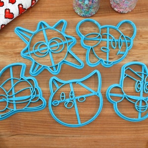 May include: Set of six blue plastic cookie cutters shaped like cartoon characters. The cutters are on a wooden surface.