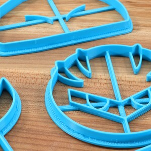 May include: A set of three blue plastic cookie cutters shaped like a spaceship, a face, and a sword. The cutters are on a wooden surface.