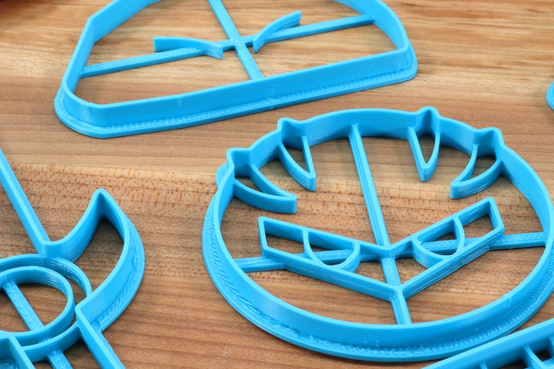 May include: A set of three blue plastic cookie cutters shaped like a spaceship, a face, and a sword. The cutters are on a wooden surface.