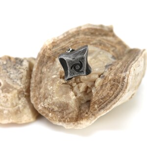 Puede incluir: Un peque&ntilde;o pendiente de metal gris oscuro con un dise&ntilde;o de espiral estilizado se muestra sobre una piedra clara y texturizada. El pendiente tiene una forma geom&eacute;trica con una apariencia ligeramente desgastada. La piedra tiene una forma natural y org&aacute;nica.