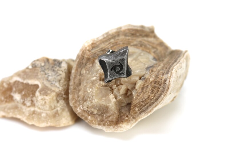 Puede incluir: Un peque&ntilde;o pendiente de metal gris oscuro con un dise&ntilde;o de espiral estilizado se muestra sobre una piedra clara y texturizada. El pendiente tiene una forma geom&eacute;trica con una apariencia ligeramente desgastada. La piedra tiene una forma natural y org&aacute;nica.