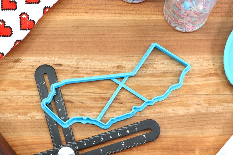 May include: A blue plastic cookie cutter shaped like the state of California. The cutter is on a wooden surface with a ruler.