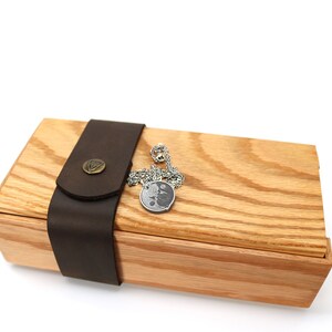 May include: A wooden box with a leather strap and a silver chain with a round pendant. The box is made of light wood with a natural grain.