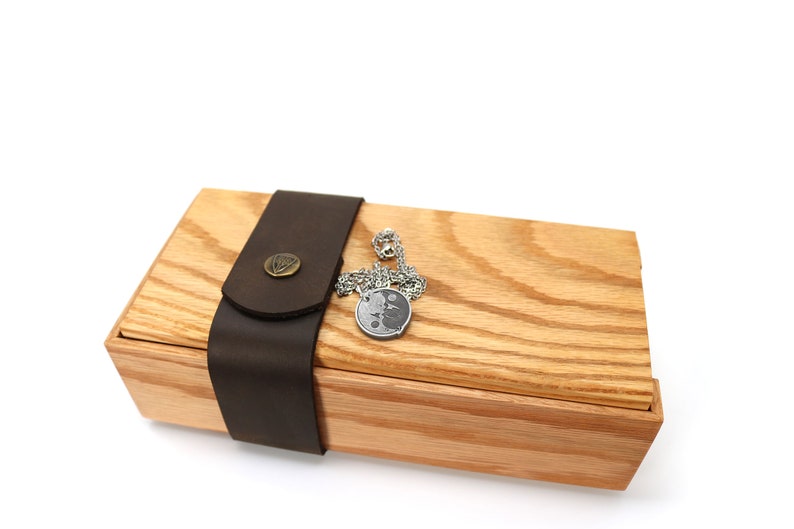 May include: A wooden box with a leather strap and a silver chain with a round pendant. The box is made of light wood with a natural grain.
