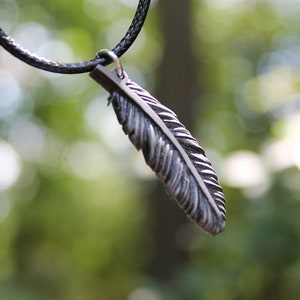 Op de afbeelding: Een zilverkleurige veervormige hanger hangt aan een zwarte koordketting. De hanger is gedetailleerd met ge&euml;tste lijnen, waardoor hij er realistisch uitziet. De achtergrond is een zacht, wazig groen, wat duidt op een buitenomgeving. Deze ketting is een sieraad.