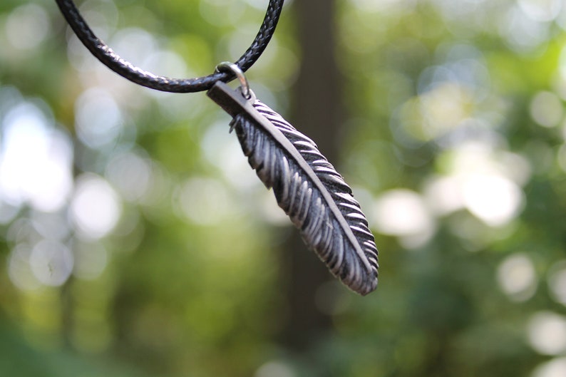 Op de afbeelding: Een zilverkleurige veervormige hanger hangt aan een zwarte koordketting. De hanger is gedetailleerd met ge&euml;tste lijnen, waardoor hij er realistisch uitziet. De achtergrond is een zacht, wazig groen, wat duidt op een buitenomgeving. Deze ketting is een sieraad.