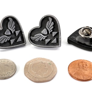 Puede incluir: Dos pines plateados en forma de coraz&oacute;n con un dise&ntilde;o de trifuerza alada. Los pines est&aacute;n al lado de un pin negro en forma de coraz&oacute;n con un cierre de pin. Tambi&eacute;n hay tres monedas en la imagen.