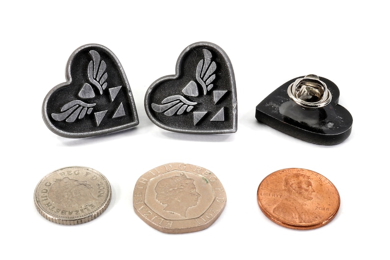 Puede incluir: Dos pines plateados en forma de coraz&oacute;n con un dise&ntilde;o de trifuerza alada. Los pines est&aacute;n al lado de un pin negro en forma de coraz&oacute;n con un cierre de pin. Tambi&eacute;n hay tres monedas en la imagen.