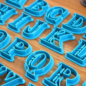 May include: A set of blue plastic alphabet cookie cutters. The cutters are arranged on a wooden surface.