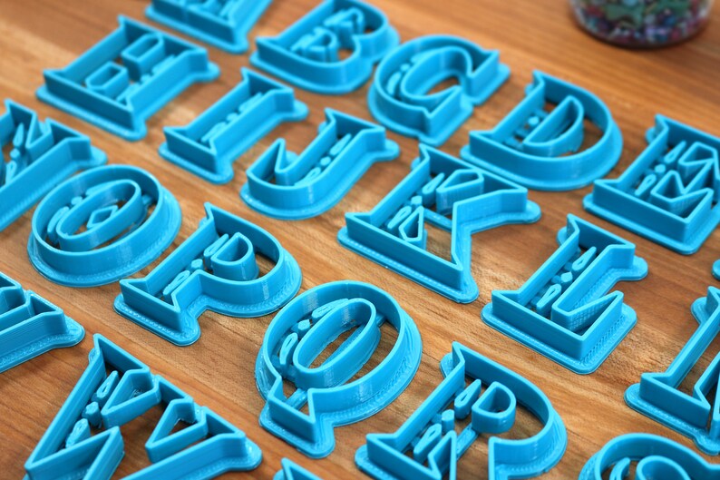 May include: A set of blue plastic alphabet cookie cutters. The cutters are arranged on a wooden surface.