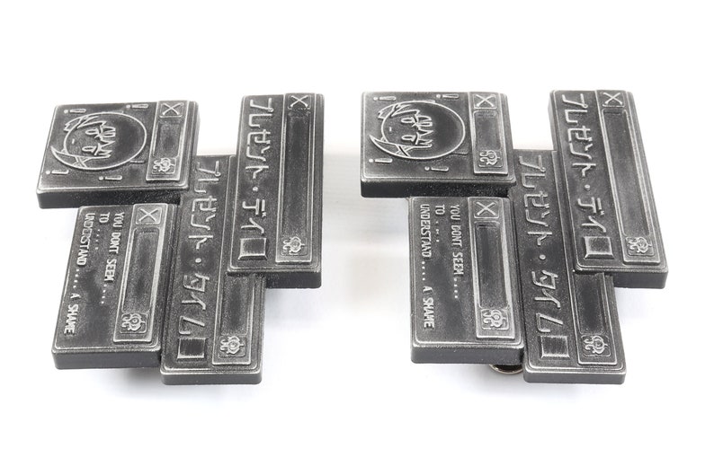 Op de afbeelding: Twee zilverkleurige metalen pinnen met een gelaagd ontwerp. Elke pin heeft een cartoonfiguur met een spraakballon met de tekst "プレゼント・タイム" en "YOU DON'T SEEM... TO... UNDERSTAND... A SHAME".