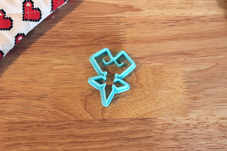 Pu&ograve; includere: Tagliapasta in plastica azzurra a forma di stella stilizzata. Il tagliapasta &egrave; su una superficie di legno, con una porzione di tessuto bianco e rosso a forma di cuore nell'angolo in alto a sinistra.