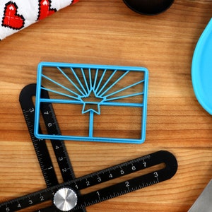 May include: A blue plastic cookie cutter shaped like the Arizona state flag. The cutter features a yellow star and red sun rays.