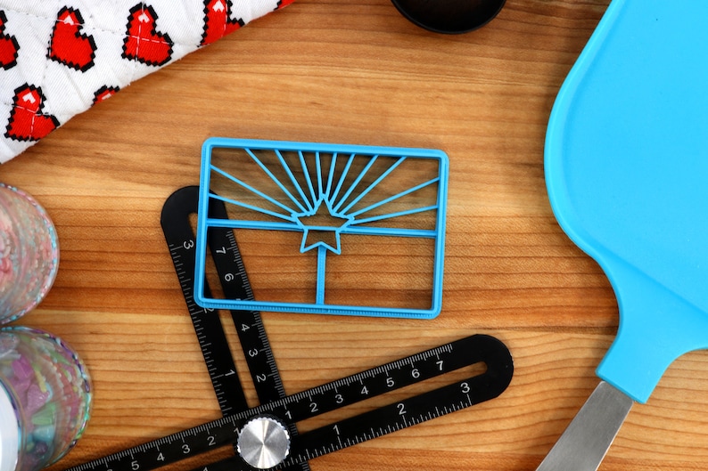 May include: A blue plastic cookie cutter shaped like the Arizona state flag. The cutter features a yellow star and red sun rays.