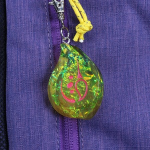 May include: A green and yellow iridescent teardrop-shaped charm with a pink symbol inside. The charm is attached to a zipper pull on a purple jacket.