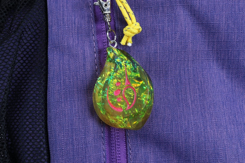 May include: A green and yellow iridescent teardrop-shaped charm with a pink symbol inside. The charm is attached to a zipper pull on a purple jacket.
