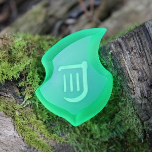 May include: A translucent green shield-shaped object with a white symbol, set on a moss-covered log. The shield has a layered design, with a lighter green centre and a darker green border. The symbol is a stylized design.