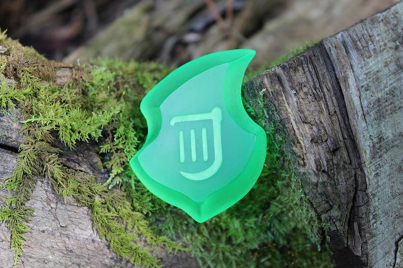 May include: A translucent green shield-shaped object with a white symbol, set on a moss-covered log. The shield has a layered design, with a lighter green centre and a darker green border. The symbol is a stylized design.
