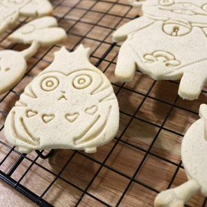May include: Five plain sugar cookies on a black wire cooling rack. The cookies are shaped like cartoon characters with various facial expressions and details. The cookies are on a wooden surface.