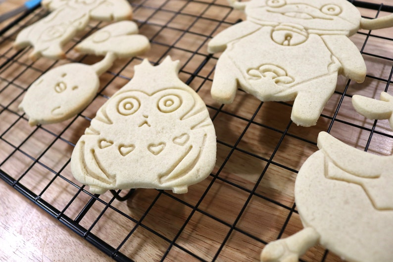 May include: Five plain sugar cookies on a black wire cooling rack. The cookies are shaped like cartoon characters with various facial expressions and details. The cookies are on a wooden surface.