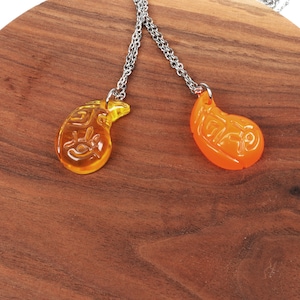 May include: Two silver chain necklaces with matching teardrop-shaped pendants. The pendants are made of clear glass with a yellow and orange colour gradient. The pendants have Chinese characters etched into them.