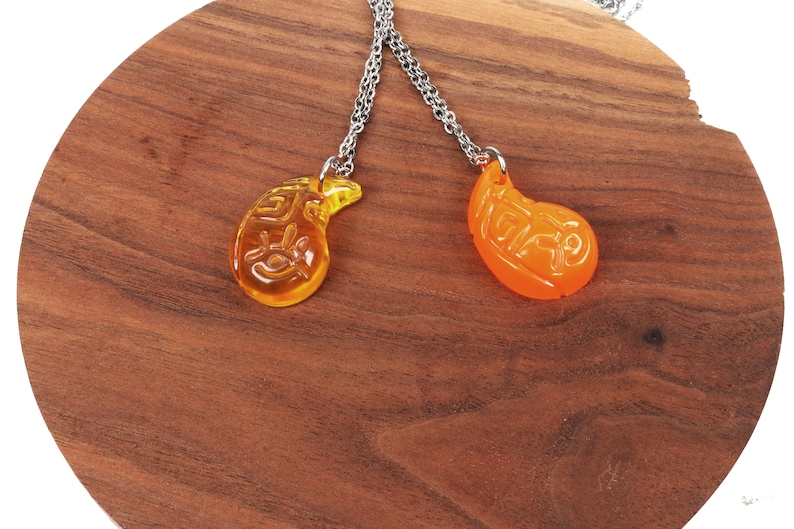 May include: Two silver chain necklaces with matching teardrop-shaped pendants. The pendants are made of clear glass with a yellow and orange colour gradient. The pendants have Chinese characters etched into them.
