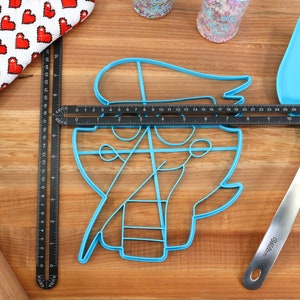 May include: A blue plastic cookie cutter shaped like a pirate with a hat, a sword, and an anchor. The cutter is on a wooden surface with a ruler and a spatula.