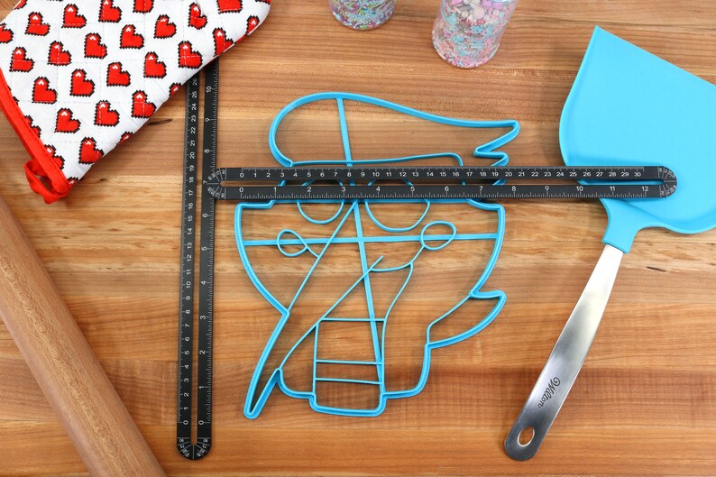 May include: A blue plastic cookie cutter shaped like a pirate with a hat, a sword, and an anchor. The cutter is on a wooden surface with a ruler and a spatula.