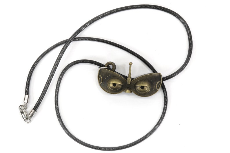 May include: A black cord necklace with a bronze pendant shaped like a robot face with large eyes and an antenna.