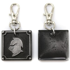 May include: A silver key chain with a black square charm featuring a knight's helmet design. The back of the charm is plain and has a worn, distressed look.