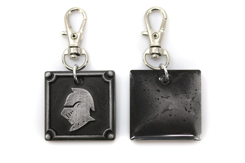 May include: A silver key chain with a black square charm featuring a knight's helmet design. The back of the charm is plain and has a worn, distressed look.