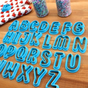 May include: A set of 26 blue plastic alphabet cookie cutters on a wooden surface. The cutters are arranged in alphabetical order.
