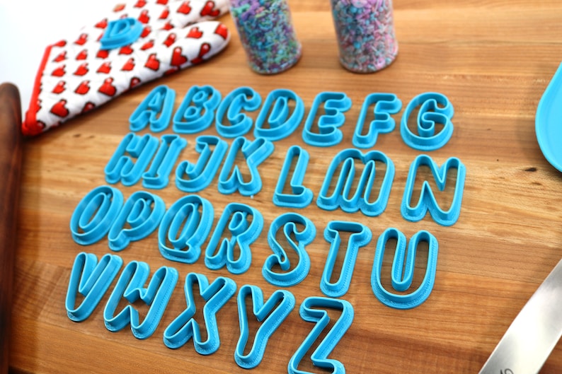 May include: A set of 26 blue plastic alphabet cookie cutters on a wooden surface. The cutters are arranged in alphabetical order.
