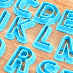 May include: A set of blue plastic alphabet cookie cutters. The cutters are arranged on a wooden surface.