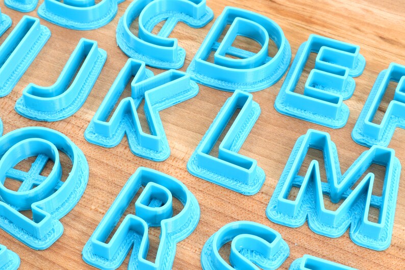 May include: A set of blue plastic alphabet cookie cutters. The cutters are arranged on a wooden surface.