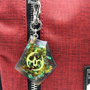 May include: A red zippered bag with a black zipper pull and a clear resin charm with gold glitter and a green and gold symbol. The charm is attached to the zipper pull.
