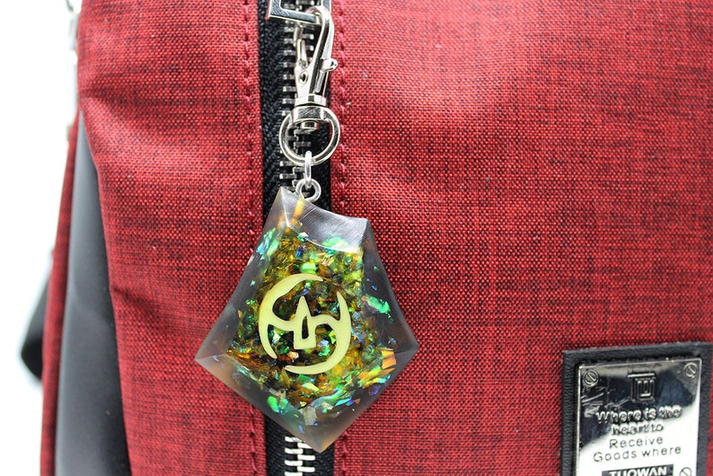 May include: A red zippered bag with a black zipper pull and a clear resin charm with gold glitter and a green and gold symbol. The charm is attached to the zipper pull.
