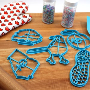 Puede incluir: Un juego de cortadores de galletas de pl&aacute;stico azul en forma de &aacute;rbol, p&aacute;jaro, cacahuete, contorno de estado con las letras "AD" dentro y un c&iacute;rculo con las letras "AD" dentro. Tambi&eacute;n hay un rodillo, una esp&aacute;tula y dos frascos de chispas.